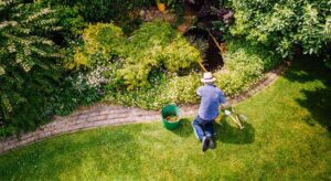 Aerial view of a person kneeling on grass weeding a large flower bed across a brick border wearing a brimmed hat