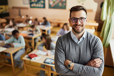 Male teacher standing in front of his classroom full of students with his arms folded