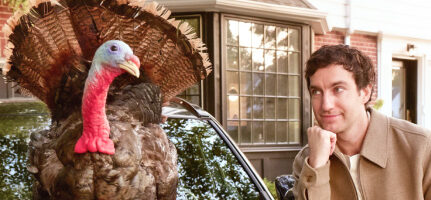 Photo of a man leaning on the hood of a car, looking at a turkey standing on the hood.
