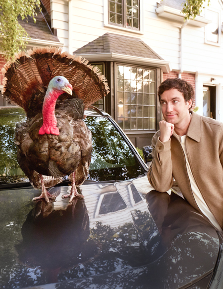 Photo of a man leaning on the hood of a car, looking at a turkey standing on the hood.