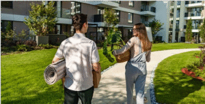 A man with a rug rolled up under his arm walks with a toman carrying a basket in a condo complex