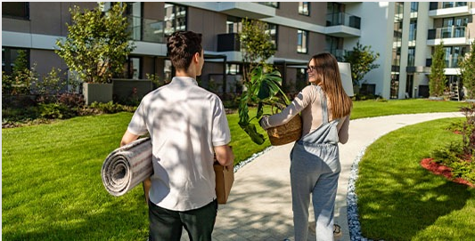 A man with a rug rolled up under his arm walks with a toman carrying a basket in a condo complex